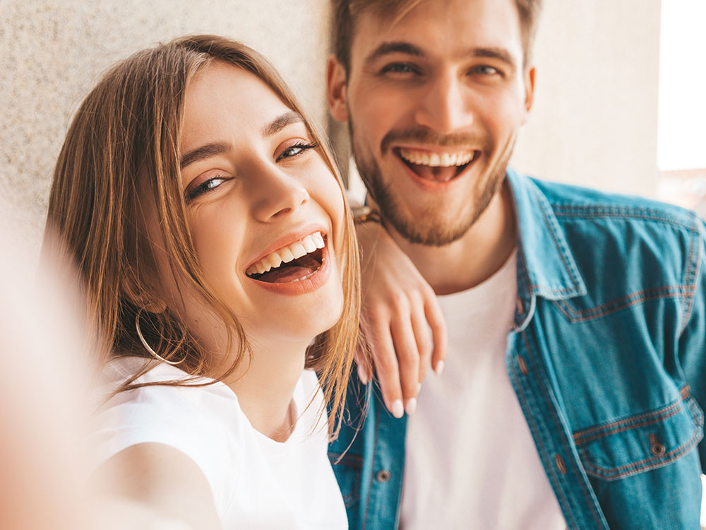 Smiling beautiful girl and her handsome boyfriend in casual summer clothes. Happy family taking selfie self portrait of themselves on smartphone camera. Having fun on the street background
